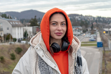 Street portrait of a young woman 25-30 years old in a hood and headphones against the background of mountains and city streets.