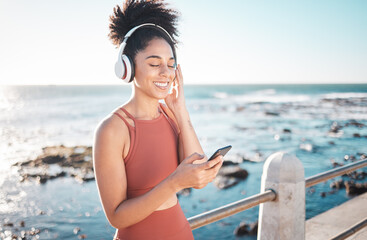 Canvas Print - Black woman at beach, headphone and smartphone with fitness, runner listening to music for sports motivation. Happy, streaming online with podcast or radio, phone and running by sea with calm outdoor