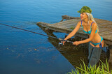 Fototapeta Tęcza - Woman fisherman in a swimsuit catches fish with a fishing rod at the pier from the shore
