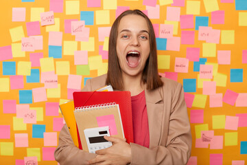 Wall Mural - Photo of amazed shocked happy woman wearing beige jacket standing against yellow wall with colorful memo cards, holding organizer and calculator, screaming happily.