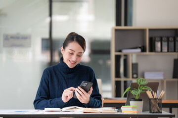 Wall Mural - Beautiful Asian woman using a smartphone and working with a laptop while sitting at an office desk, working from home concept.