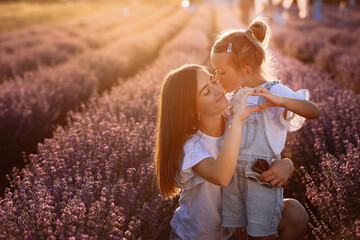 Wall Mural - Mothers day. Young mom with little child daughter doing gesture heart with hands, hugging and kissing in a blooming lavender field. Family of two having fun, playing in meadow field on summer holiday