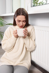 Poster - Woman holding cup with hot drink near heating radiator indoors