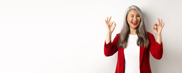 Asian professional businesswoman showing OK signs and winking, smiling pleased, assure or recommend something, standing against white background