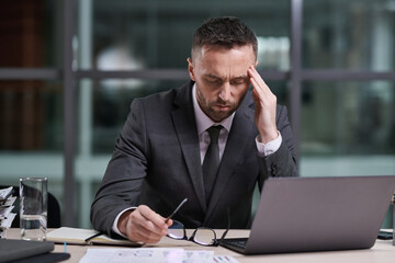 Poster - Stressed male director of business corporation touching head by hand and bending over workplace with laptop while working overtime in office