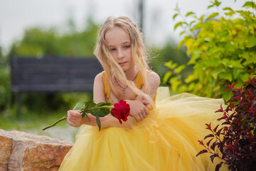 Wall Mural - A little girl with blond hair in a lush yellow dress walks in the park with a red rose. Princess image.