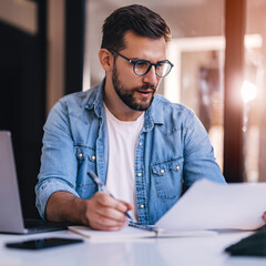 Satisfied young man with glasses sitting at a desk and doing paperwork at his workplace.