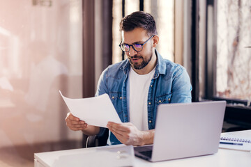 Wall Mural - Satisfied young man with glasses sitting at a desk and doing paperwork at his workplace.