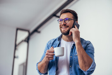 Handsome bearded man leaning on office desk, drinking coffee and talking on the phone.