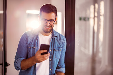 Wall Mural - Handsome young man with glasses standing in the office and using the phone.