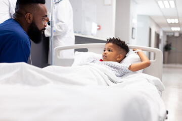 African american male doctor talking to boy patient in hospital bed in corridor with copy space