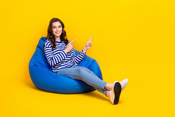 Poster - Full length portrait of cheerful nice person sit comfy bag indicate fingers empty space isolated on yellow color background