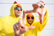 Friends Wearing Yellow Chicken Costumes Looking Through Finger Frame In Front Of Wall