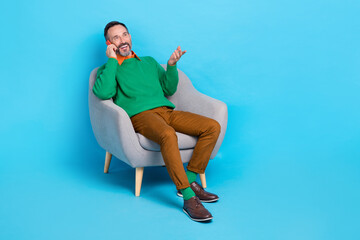 Sticker - Full body photo of mature handsome man sitting armchair talking phone roaming wear trendy green garment isolated on blue color background