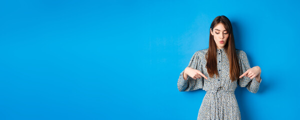 Intrigued beautiful girl in dress say wow, looking and pointing down at advertisement, showing promo with interested face, blue background