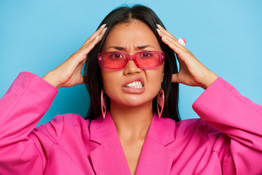 Portrait of young Asian woman, dressed in fashion pink jacket and glasses, isolated by blue background feeling frustrated with helpless face expression