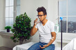 Portrait of a sick young african american man in the glasses sitting in a white medical ward under a dropper. Male sad patient on the bed drinking a glass of water. Healthcare and medicine concept.