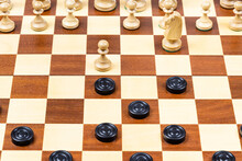 Playing By Different Rules On The Same Board - Black Checkers And White Chess Figures On Wooden Chessboard, Beginning Of Chess Checkers Game (focus On Center Of Board)