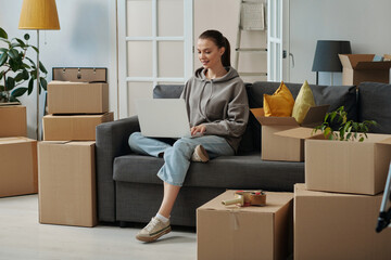 Girl sitting on sofa and using laptop to search for movers during her moving in new house