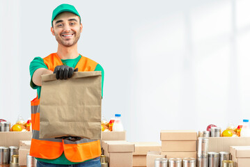 Wall Mural - Help Collection Center, Free Food Distribution. Volunteer carrying food donation box. Young smiling man wearing green uniform cap and t-shirt, orange vest holds out grocery set for in-need people