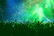© tsuguliev - A crowded concert hall with scene stage green lights, rock show performance, with people silhouette, colourful confetti explosion fired on dance floor air during a concert festival
