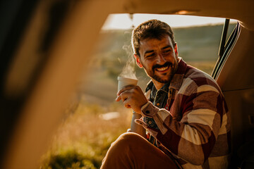 Handsome man drinking coffee while sitting in a car and enjoying the sunset.