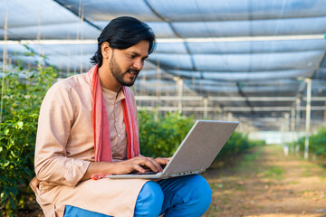 Happy young farmer busy working on laptop while sitting at greenhouse - concept of modern farming, technology and development or growth.