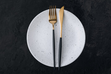 Empty plate with fork and knife on dark stone background. Gold and black tableware with white plate