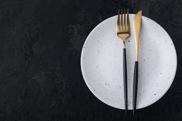 Empty plate with fork and knife on dark stone background. Gold and black tableware with white plate