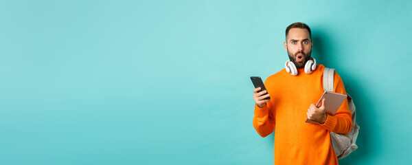 Wall Mural - Handsome man student with headphones and backpack, holding digital tablet and smartphone, staring confused at camera, standing against turquoise background
