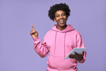 Wall Mural - Happy smiling African American teen boy student wearing pink hoodie holding using digital tablet pointing finger aside at copy space advertising, presenting isolated on light purple background.