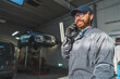 © PoppyPix - A car mechanic in a grey jacket at an auto repair shop with lifted cars for repair. High-quality photo