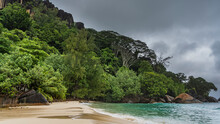 Tropical Landscape. The Foam Of The Waves Of The Turquoise Ocean On The Beach. Wet Sand Glistens. A Hill Overgrown With Green Vegetation, Against A Cloudy Sky. Boulders At The Water's Edge. Seychelles