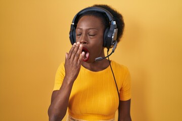 Wall Mural - African woman with curly hair standing over yellow background wearing headphones bored yawning tired covering mouth with hand. restless and sleepiness.