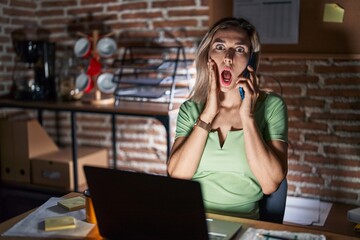 Wall Mural - Young beautiful woman working at the office at night speaking on the phone afraid and shocked, surprise and amazed expression with hands on face