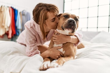 Poster - Young caucasian woman hugging and kissing dog lying on bed at bedroom