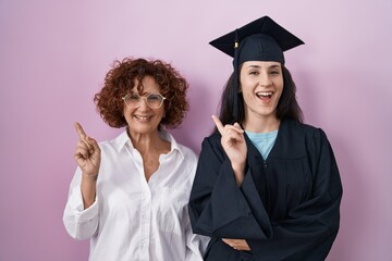 Sticker - Hispanic mother and daughter wearing graduation cap and ceremony robe with a big smile on face, pointing with hand and finger to the side looking at the camera.