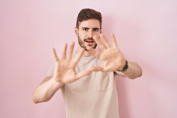 Hispanic man with beard standing over pink background afraid and terrified with fear expression stop gesture with hands, shouting in shock. panic concept.