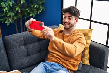 Canvas Print - Young hispanic man make selfie by smartphone sitting on sofa at home