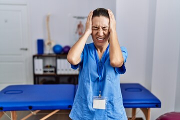 Wall Mural - Young hispanic woman wearing physiotherapist uniform standing at clinic suffering from headache desperate and stressed because pain and migraine. hands on head.