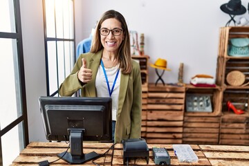 Poster - Young hispanic woman working as manager at retail boutique smiling happy and positive, thumb up doing excellent and approval sign