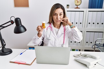 Wall Mural - Young hispanic woman wearing doctor uniform holding pills at the clinic mouth and lips shut as zip with fingers. secret and silent, taboo talking