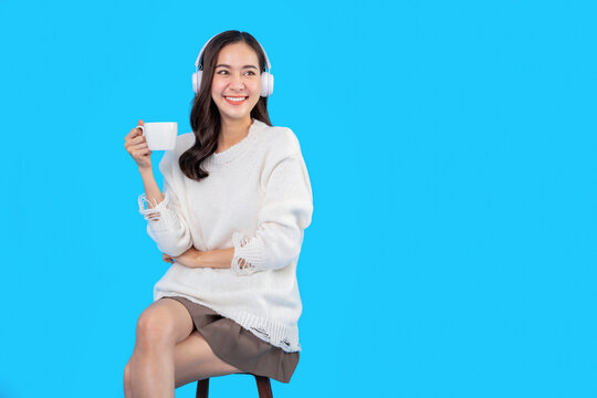 Fototapete - Young asian woman sitting in chair, holding coffee cup in hands, enjoying her free time from work, isolated on light blue background
