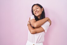 Young Hispanic Woman Standing Over Pink Background Hugging Oneself Happy And Positive, Smiling Confident. Self Love And Self Care