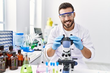 Sticker - Young hispanic man wearing scientist uniform using microscope at laboratory