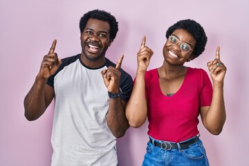 Sticker - Young african american couple standing over pink background smiling amazed and surprised and pointing up with fingers and raised arms.