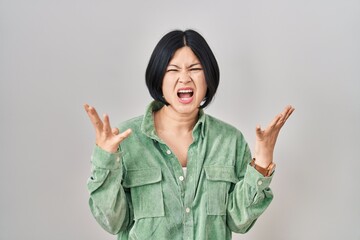 Wall Mural - Young asian woman standing over white background crazy and mad shouting and yelling with aggressive expression and arms raised. frustration concept.