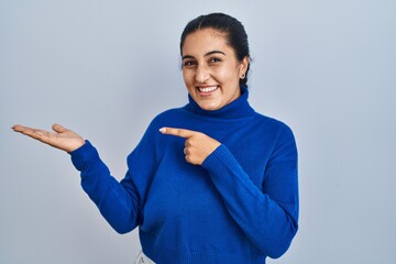 Poster - Young hispanic woman standing over isolated background amazed and smiling to the camera while presenting with hand and pointing with finger.