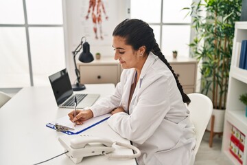 Poster - Young hispanic woman wearing doctor uniform writing medical report at clinic