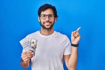 Poster - Handsome latin man holding dollars banknotes smiling happy pointing with hand and finger to the side